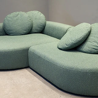 Sofá modular verde, com pés de madeira, almofadas de assento e encosto fixas e quatro almofadas decorativas, visto de lado em um fundo de parede cinza.