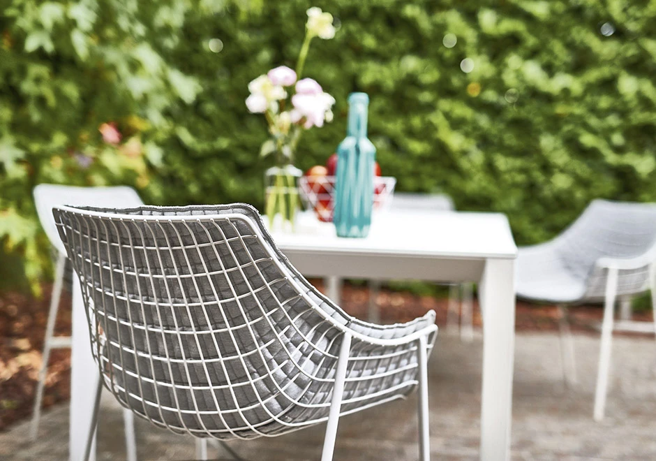 Cadeira de design com estrutura de aço cromado ou pintado e tecido branco, vista de costas, em uma area externa.