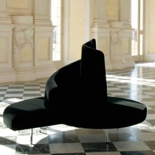 Sofá ilha,com pés em aço cromado e estofado de veludo preto, visto de frente em um salão vazio.