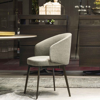 Cadeira assinada estofada cinza com pés escuros vista de lado, sobre um tapete claro, com grandes móveis de madeira ao fundo e um objeto dourado do lado esquerdo.