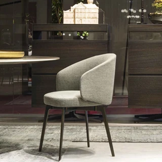 Cadeira assinada estofada cinza com pés escuros vista de lado, sobre um tapete claro, com grandes móveis de madeira ao fundo e um objeto dourado do lado esquerdo.