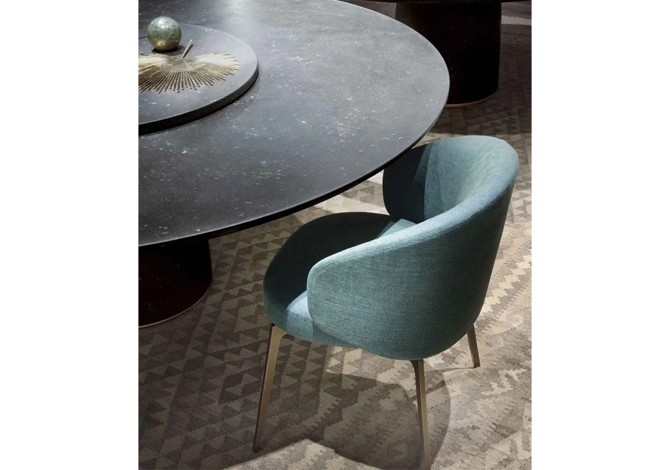 cadeira assinada estofada azul com pés escuros vista de cima ao lado de uma mesa redonda preta não aparece por completo sobre um tapete com estampa de triângulos.