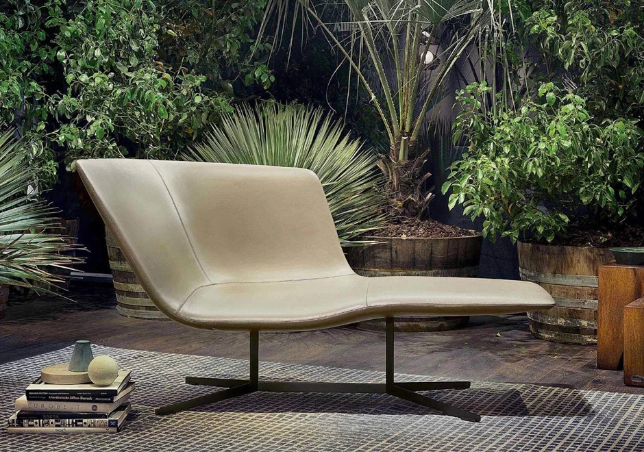 Chaise-longue com estrutura de aço com acabamento bronze, revestida em espuma moldada em diferentes densidades, estofada em couro bege, visto de lado em uma area externa.