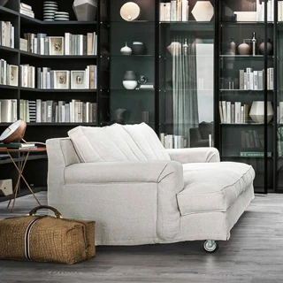 sofá aconchegante de três lugares visto de lado, com uma maleta ao lado e uma grande estante ao fundo, cheia de livros e outros objetos, vasos com plantas do lado direito.