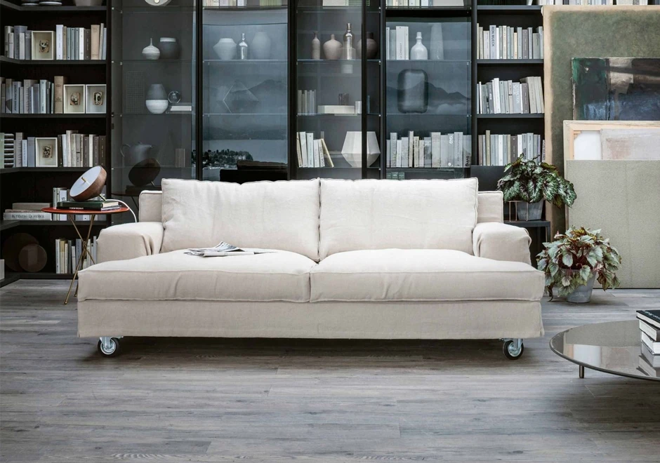 sofá aconchegante de três lugares visto de frente, com uma grande estante ao fundo, cheia de livros e outros objetos, vasos com plantas e quadros sobrepostos do lado direito.