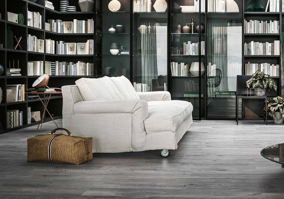 sofá aconchegante de três lugares visto de lado, com uma maleta ao lado e uma grande estante ao fundo, cheia de livros e outros objetos, vasos com plantas do lado direito.