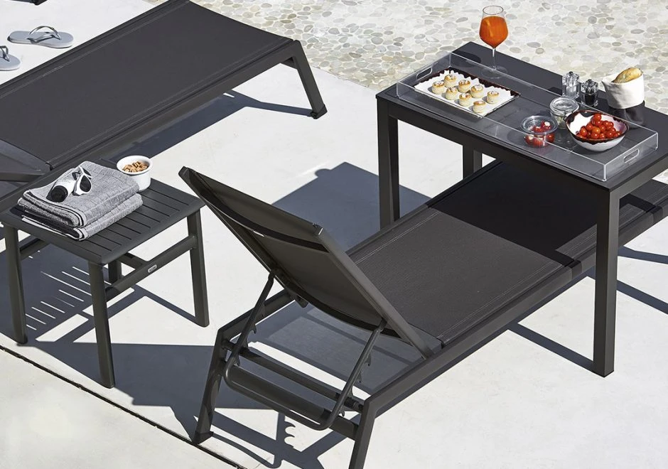 Chaise longue com estrutura de alumínio, assento e encosto em PVC preto, visto de costas em uma area externa.